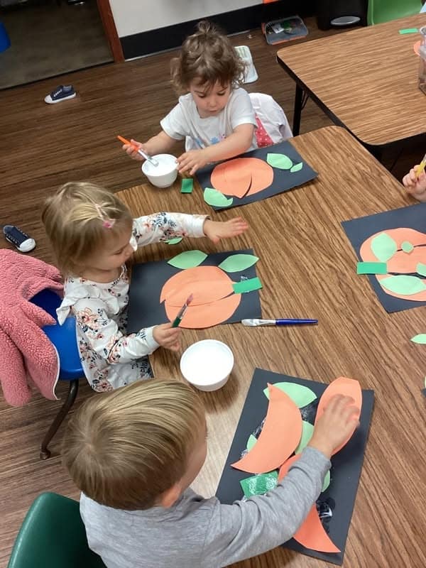 Three young children sitting at a table working on paper peach craft projects with glue and brushes.