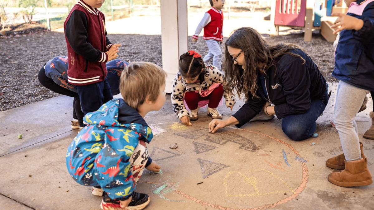 An adult woman kneels on a patio to draw with chalk alongside four children.