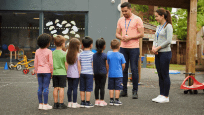 Two adults stand facing a group of young children lined up outside near playground equipment at a daycare or school.